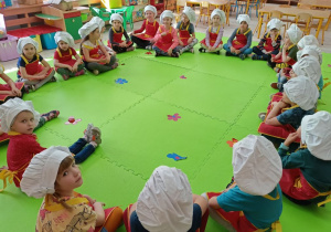 Dzieci ubrane w stroje kucharskie siedzą w kole na dywanie.